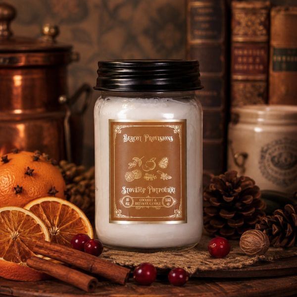 A white wax candle in a clear glass jar with a black lid and a brown label reading Stovetop Potpourri styled on a rustic wooden surface with dried orange slices, cinnamon sticks, red berries, pinecones, and old books in the background.