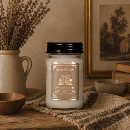 Candle with a decorative label on a rustic surface with a jar of lavender and framed picture in the background.