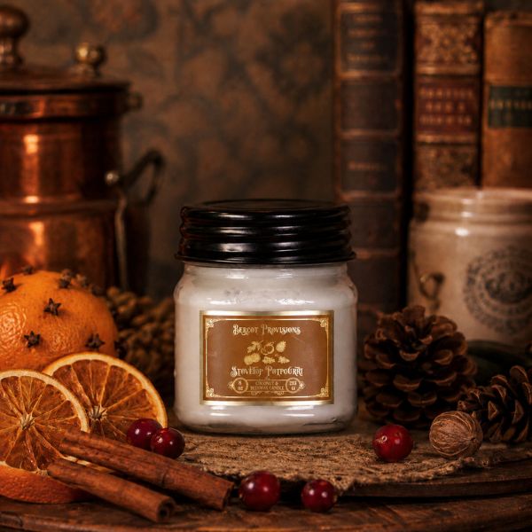 A white wax candle in a clear glass jar with a black lid and a brown label reading Stovetop Potpourri styled on a rustic wooden surface with dried orange slices, cinnamon sticks, red berries, pinecones, and old books in the background.