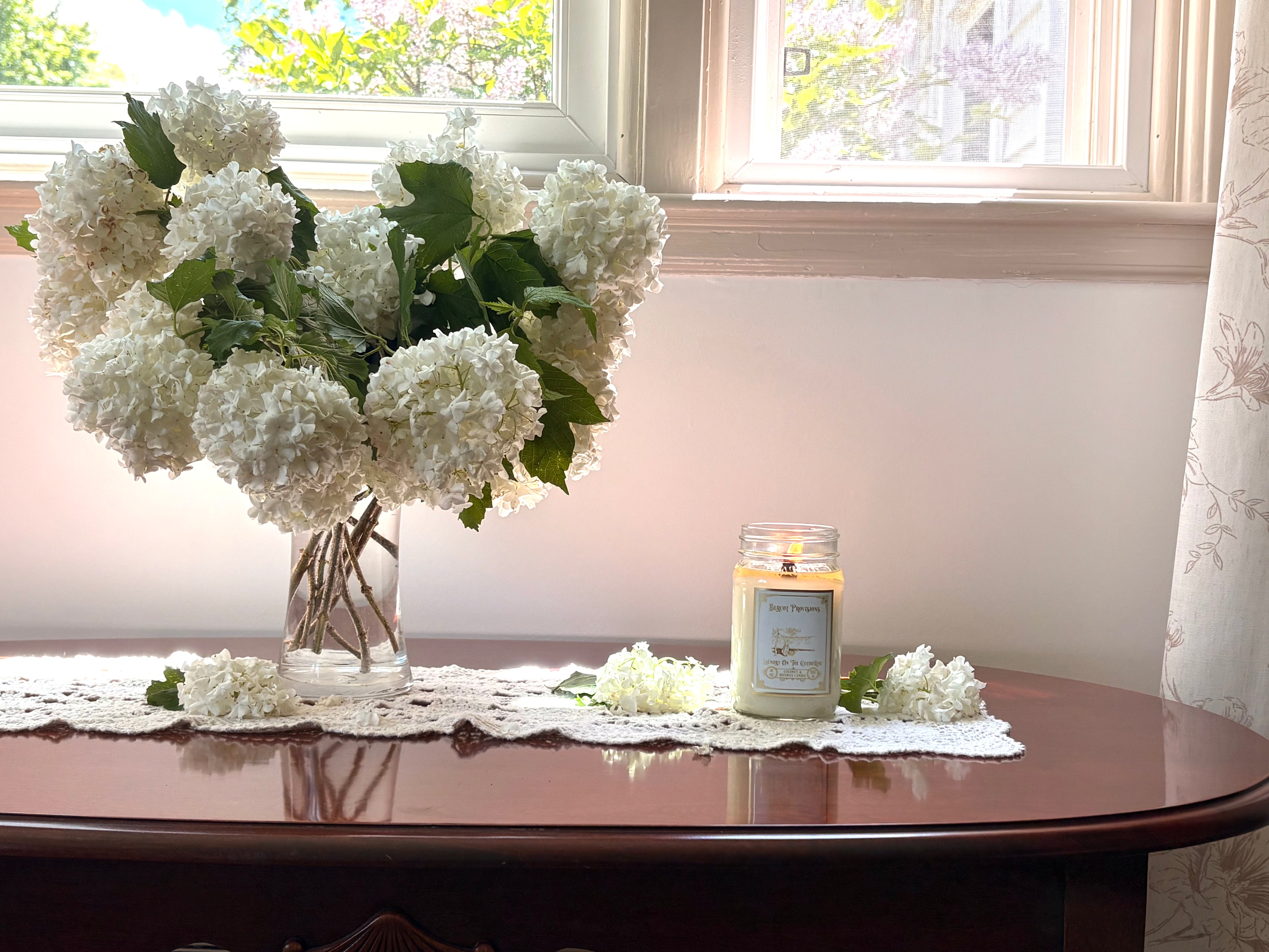 White flowers in a vase and Laundry on the Clotheline candle lit on a wooden table with a window in the background.