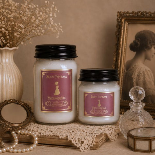 Two candles with labels on a decorative surface with a vintage frame and vase in the background.