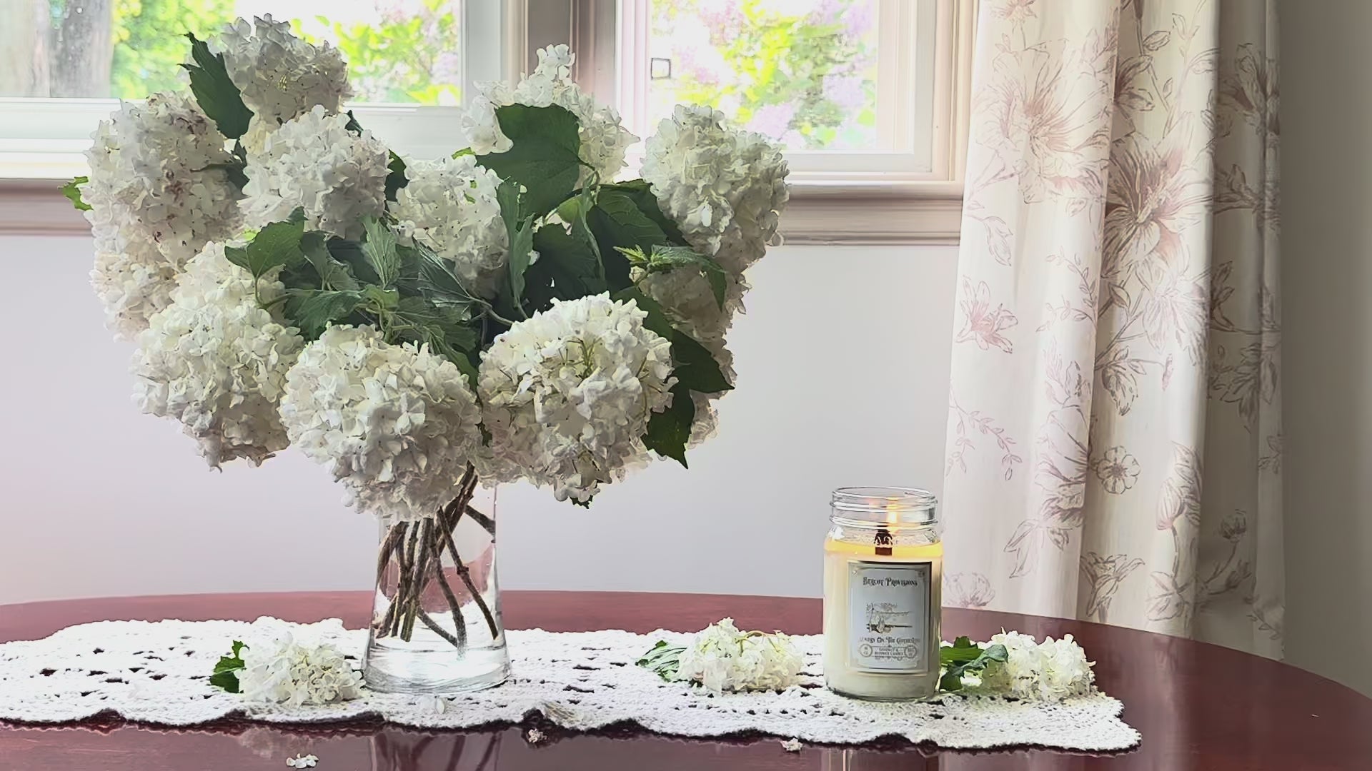 Load video: Laundry on the Clothesline candle burning on a sunny afternoon on the coffee table next to a vase full of white flowers.